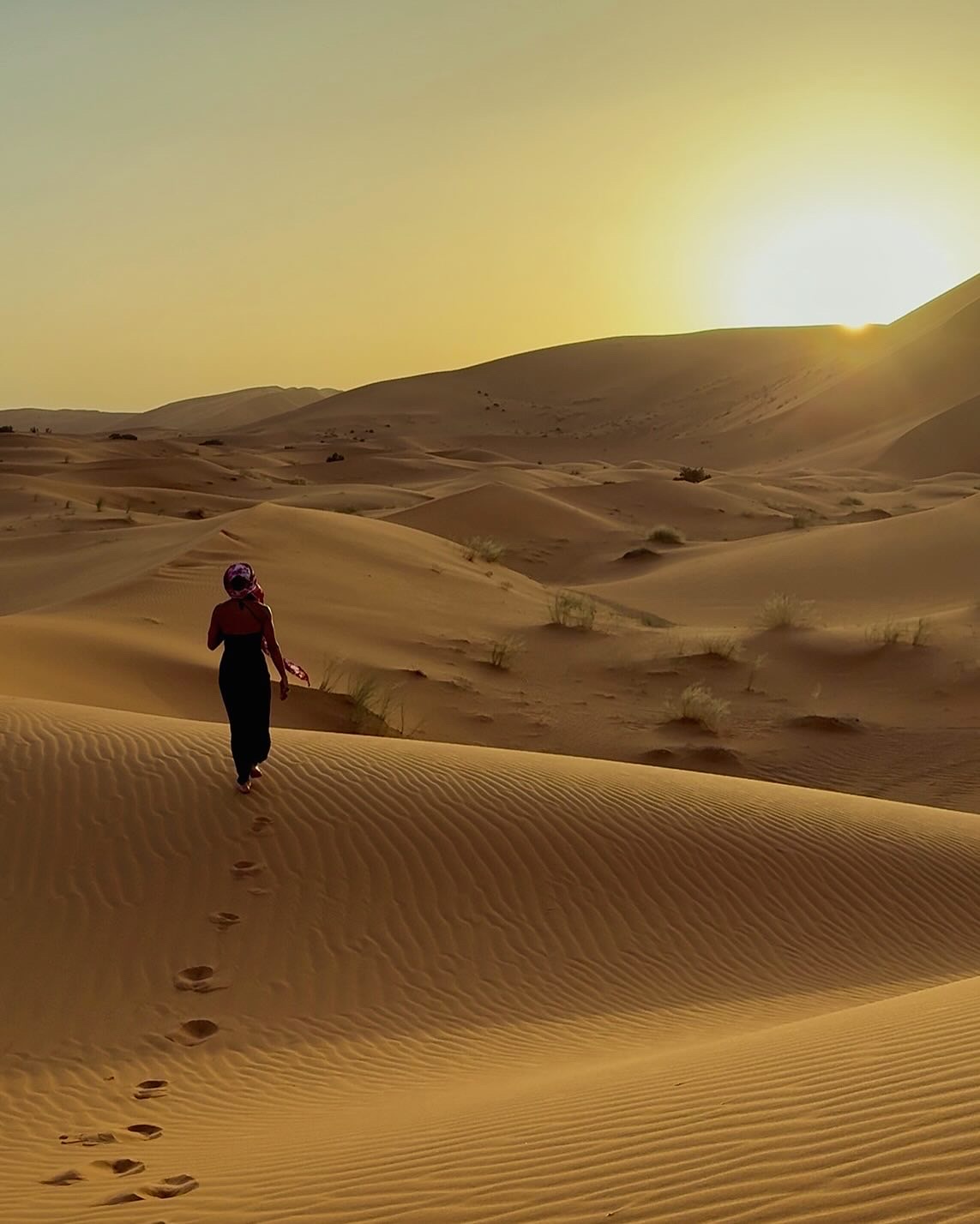 A woman walking across Sahara dunes toward the setting sun, leaving a trail of footprints in the sand